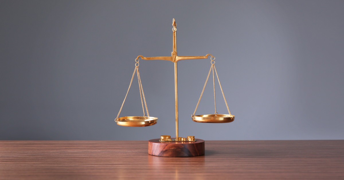 A small brass balance scale sits on a light wooden table, with a blurred laptop and documents in the background.